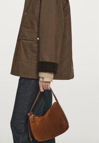 Braune Wildleder-Handtasche mit geschwungener Form und einem Griff, in einer Hand gehalten. Kombiniert mit einer braunen Außenjacke, einem beigen Pullover und dunklen Jeans.