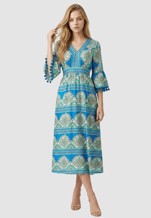 Mujer con un vestido midi azul con estampado, mangas tres cuartos tipo campana y ribete de pompones, de pie con una mano tocándose el cabello.