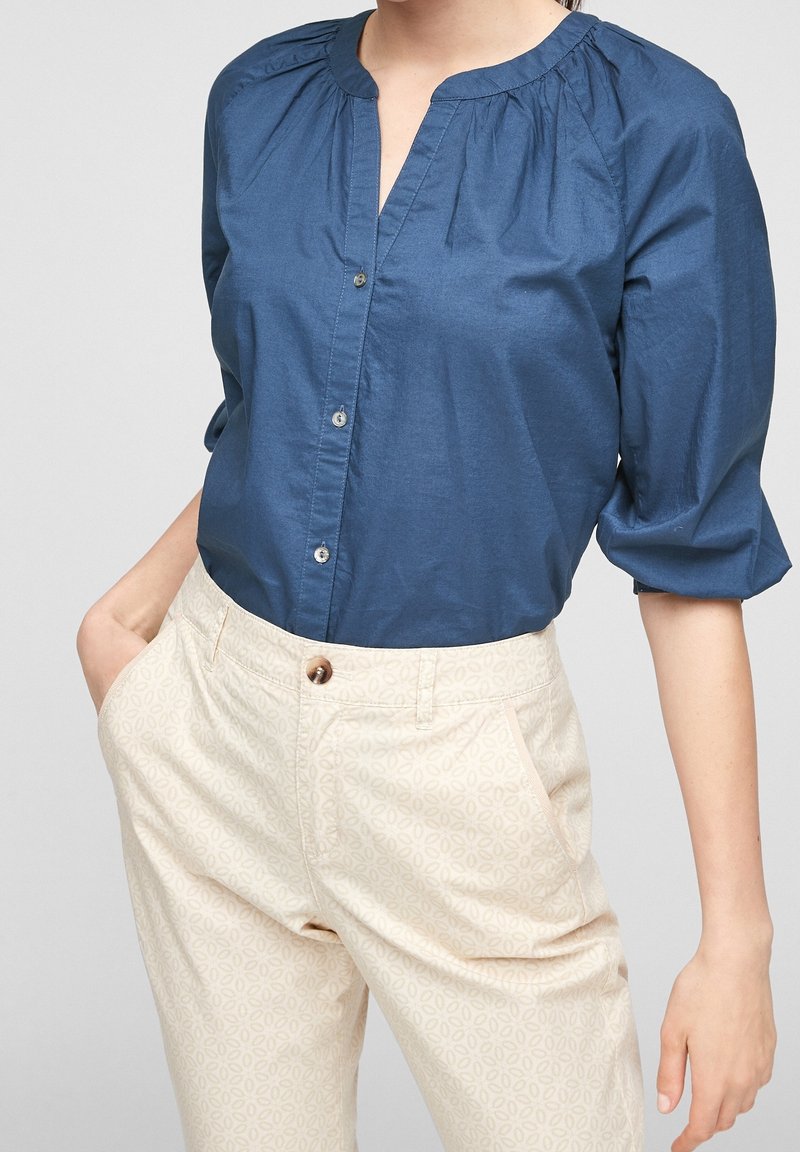 Mujer vistiendo una blusa azul con botones y mangas hasta el codo, y pantalones crema con un patrón, con las manos en los bolsillos, frente a un fondo liso.