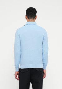 Rövid, göndör hajú férfi világoskék, bordázott pulóverben és fekete nadrágban, háttal állva, egyszínű fehér háttér előtt.