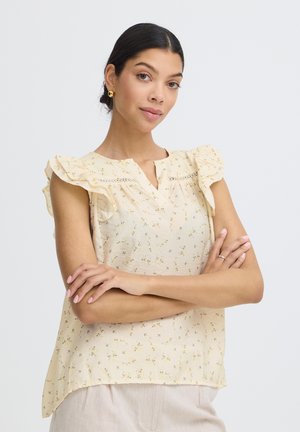 Mujer con el cabello oscuro recogido en un moño, lleva pendientes dorados y una blusa sin mangas con volantes y estampado floral en amarillo claro, con los brazos cruzados, sobre un fondo liso.