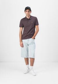 Dunkelbraunes Poloshirt mit gelbem Logo, hellblaue Jeansshorts, weiße Sneakers und schwarze Kappe, vor einem einfarbigen Hintergrund stehend.