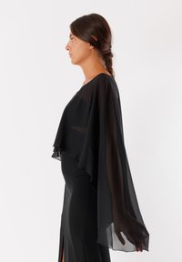 Top in chiffon nero a cappotto con maniche ampie, design a strati e orlo spaccato, indossato sopra un abito aderente nero con uno spacco sulla gamba.