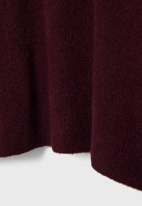 Tissu tricoté doux de couleur bordeaux foncé avec une texture visible et un ourlet côtelé, drapé sur un fond clair uni.