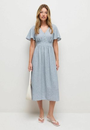 Young woman wearing a light blue midi dress with flutter sleeves, holding a white handbag and wearing white heeled sandals, standing on a plain background.