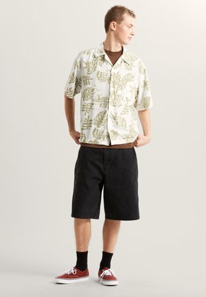 Camisa blanca de manga corta con estampado de hojas verdes, combinada con pantalones cortos negros. Zapatos de lona rojos con suelas blancas. Calcetines negros visibles.