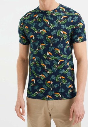 Mand iført en marineblå kortærmet skjorte med et farverigt tukan- og grønne bladmønstre, kombineret med lysebrune bukser.