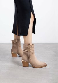 Botines de ante beige con un cuello doblado, detalle de borlas y un tacón marrón apilado con acentos dorados. Textura suave.