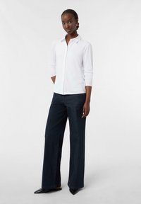 Camicia bianca con bottoni a maniche lunghe abbinata a jeans a gamba larga blu scuro. L'outfit presenta un collo a camicia e scarpe a punta.