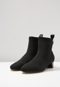 Bottines noires en tissu tricot extensible, avec un bout pointu et un petit talon carré. Dotées d'une tige côtelée à la cheville et d'une languette à l'arrière pour faciliter l'enfilage.