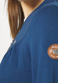 Marinblå tröja med strukturerad yta, med en rund läderlapp på ärmen och metalldekorationer. Mjukt tyg med veckdetaljer.