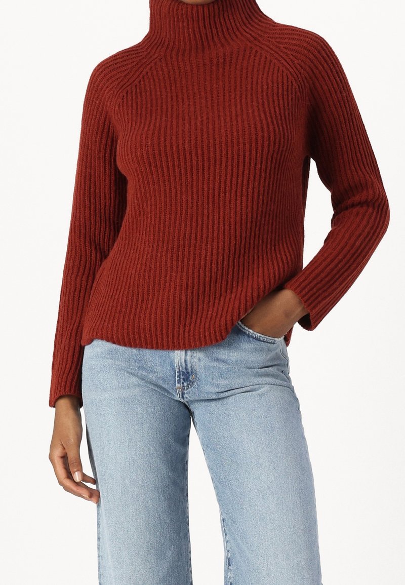 Personne portant un pull à col roulé côtelé couleur rouille et un jean bleu clair, une main dans la poche et l'autre détendue sur le côté.