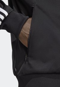 Hand i svart dragkedjeficka på svart träningsjacka med ribbad mudd och vita ränder på ärmen.