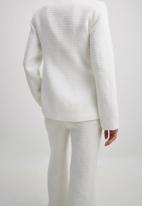 Bílý texturovaný oblek s fitted sakem a širokými nohavicemi. Tkanina má jemný tkaný vzor a hladký povrch.
