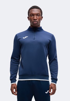 Jeune homme debout, portant un survêtement de sport bleu marine avec un logo blanc sur la poitrine et le pantalon, fond clair uni.