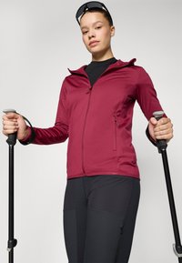 Weinroter Reißverschlussjacke aus glattem Stoff, ausgestattet mit zwei seitlichen Reißverschlusstaschen, kombiniert mit schwarzen enganliegenden Hosen und Wanderstöcken.