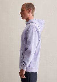 Felpa viola chiaro in misto cotone, con tasca frontale, polsini a costine e cappuccio con cordino, indossata da una persona di profilo.