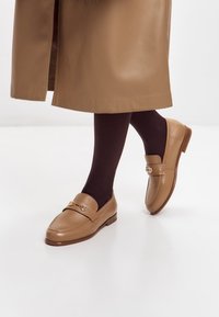 Hnědé kožené mokasíny mají zaoblenou špičku, dekorativní pásek se kovovou sponou a nízký plochý podpatek, doplněné tmavými punčochami.