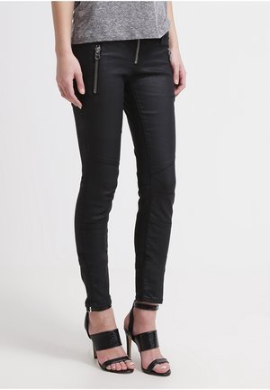 Žena oblečená v černých přiléhavých kalhotách s kapsami na zip a černých sandálech na podpatku, stojící před jednoduchým světlým pozadím.