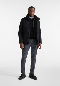 Giacca impermeabile nera con colletto alto, abbinata a un maglione lavorato a maglia blu navy e pantaloni a quadri grigi. Sneakers nere completano l'outfit.