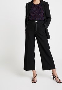 Black tailored trousers with wide legs, white stitching details, a front button closure, paired with a dark patterned top and blazer.