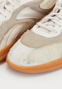 Adidași bej și albi, cu un amestec de piele întoarsă și materiale textile, talpă din cauciuc și detalii de cusătură ușoară.