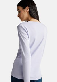 Eine Frau mit schokobraunem Haar trägt ein helllila Langarmshirt, blickt seitlich mit leicht gesenktem Kopf vor einem schlichten weißen Hintergrund.