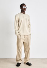 Polo de manga longa bege com colarinho abotoado, combinado com calças largas bege e sapatos pretos. O modelo usa óculos de sol espelhados.