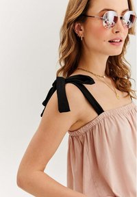 Žena v ružovom bezrúškovom topu s čiernymi šnúrkami na ramienkach, okrúhlymi reflektujúcimi slnečnými okuliarmi, zlatými náhrdelníkmi a malými zlatými kruhovými náušnicami.