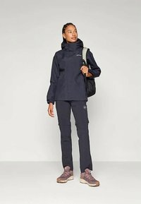 Kvinne iført mørk marineblå utendørsjakke og bukser, bærer en svart rygsekk, står med hodet vendt litt til høyre, lyse joggesko.