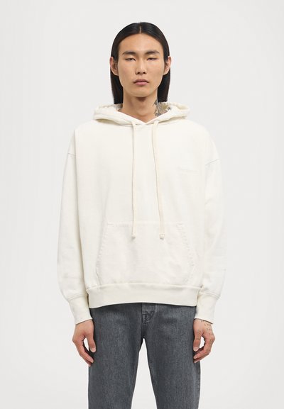 Light-colored hoodie made of soft fabric, featuring a front kangaroo pocket, drawstrings, and ribbed hem. Worn with dark grey jeans.
