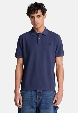Ung mand med krøllet mørkt hår iført en navyblå kortærmet poloshirt og blå jeans, stående foran en ensfarvet lys baggrund.