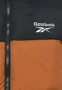 Padded jakna u crnoj i smeđoj boji s patentnim zatvaračem sprijeda. Ima bijeli Reebok logo na prsima i glatku teksturu. Uključeni su reflektirajući detalji.