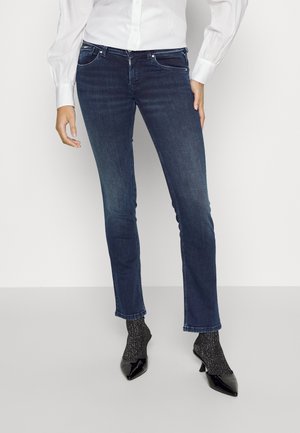 Person iført mørkeblå skinny jeans, en hvid skjorte med knapper, sorte glitrende sokker og sorte spidse hæle, der står på et almindeligt gulv.