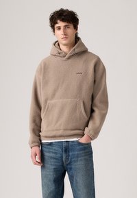 Beige fleece hoodie with a kangaroo pocket, drawstring hood, and "LEVI'S" logo. Paired with blue jeans. Soft, textured material.