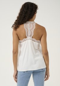 White sleeveless top featuring a sheer lace back, with scalloped edges and a smooth front. Paired with light blue jeans.