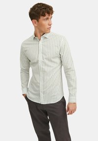 Chemise blanche à boutons avec motif à pois noirs et bleus, manches longues et coupe ajustée. Portée avec un pantalon sombre.
