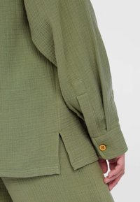 Grünes, strukturiertes Baumwollhemd mit Knopfverschluss, seitlichen Schlitzen und einer lockeren Passform. Der Stoff hat ein weiches, zerknittertes Aussehen.