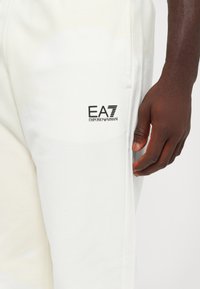 White joggers made of soft fabric, featuring a black EA7 Emporio Armani logo on the left thigh, with a relaxed fit and subtle texture.