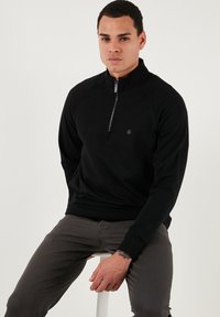 Melna zip-up vilnas krekla ar augstu apkakli, ribotām piedurknēm un vienkāršu logotipu uz krūšu daļas, apvienota ar pelēkiem šauriem biksēm.