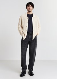Uomo in piedi con cardigan beige abbottonato, t-shirt nera, jeans neri sbiaditi, scarpe in pelle nere, mani nelle tasche del cardigan, sfondo bianco.