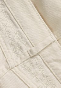 Tissu couleur crème avec des broderies complexes de motifs de feuilles. Des coutures plates et un petit détail en boucle rehaussent le design. Texture douce.