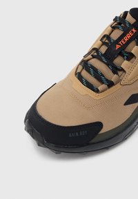 Barna szintetikus cipő fekete gumitalppal, texturált felsőrésszel, fekete fűzőkkel kék részletekkel, valamint "RAIN.RDY" felirattal az oldalon.