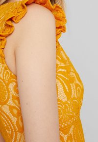 Gul spetsplagg med volangkrage och intrikata blommönster, som visar en texturerad tyg och en figursydd design.