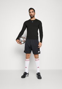 Schwarzes Langarmshirt, graue Shorts und weiß-rot gestreifte Socken. Schwarze Fußballschuhe. Hält einen schwarz-weißen gemusterten Fußball.