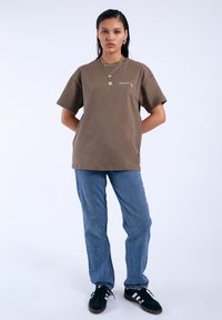 Camiseta de algodón marrón de manga corta con un pequeño logo. Combinada con unos vaqueros de mezclilla azules y zapatillas negras con acentos blancos.