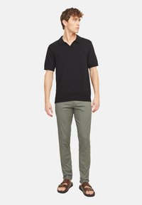 Polo shirt noir à manches courtes avec une texture douce, associé à un pantalon slim-fit olive vert et des sandales marron, se tenant devant un fond blanc.