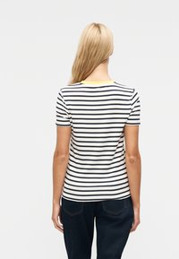 Ξ ανθιά γυναίκα που φοράει λευκό t-shirt με μαύρες οριζόντιες ρίγες και σκούρο μπλε τζιν, όρθια με την πλάτη προς το μέρος μας σε απλό λευκό φόντο.