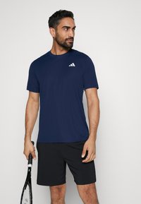 Camiseta deportiva de color azul marino hecha de tejido transpirable, con cuello redondo, mangas cortas y un logo blanco en el lado izquierdo del pecho.