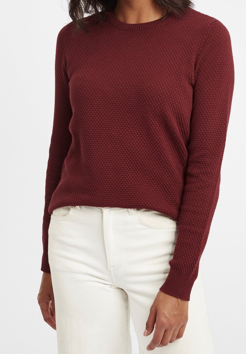 Pull texturé bordeaux à col rond et manches longues, associé à un jean de couleur claire. Motif en maille détaillé visible sur le tissu.